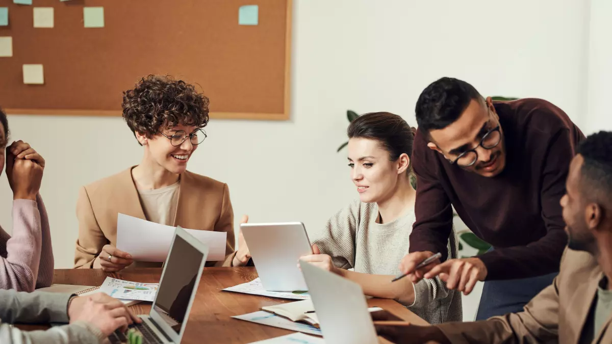 A group of people are sitting around a table in a meeting room. They are all looking at a laptop computer. The room is well-lit and the people are dressed professionally.