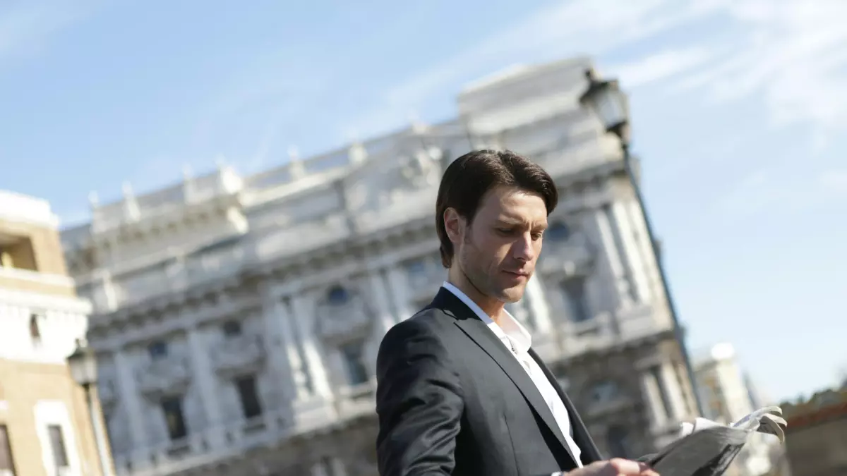 A man in a suit stands in front of a large building, holding a newspaper.