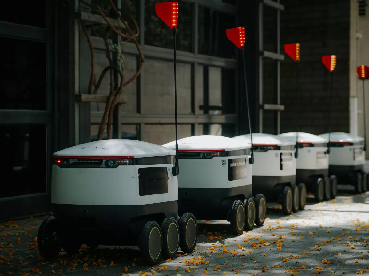 A row of delivery robots lined up against a building. They are white with black accents, and have a rounded shape. They are all facing the same direction, with their headlights on.