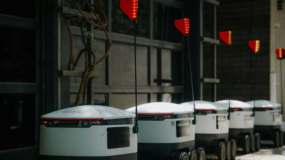 A row of delivery robots lined up against a building. They are white with black accents, and have a rounded shape. They are all facing the same direction, with their headlights on.