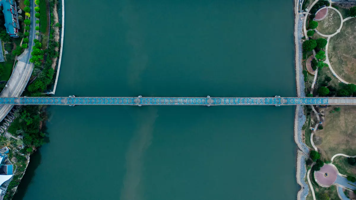 An aerial view of a bridge crossing a river, with green trees and buildings in the background.