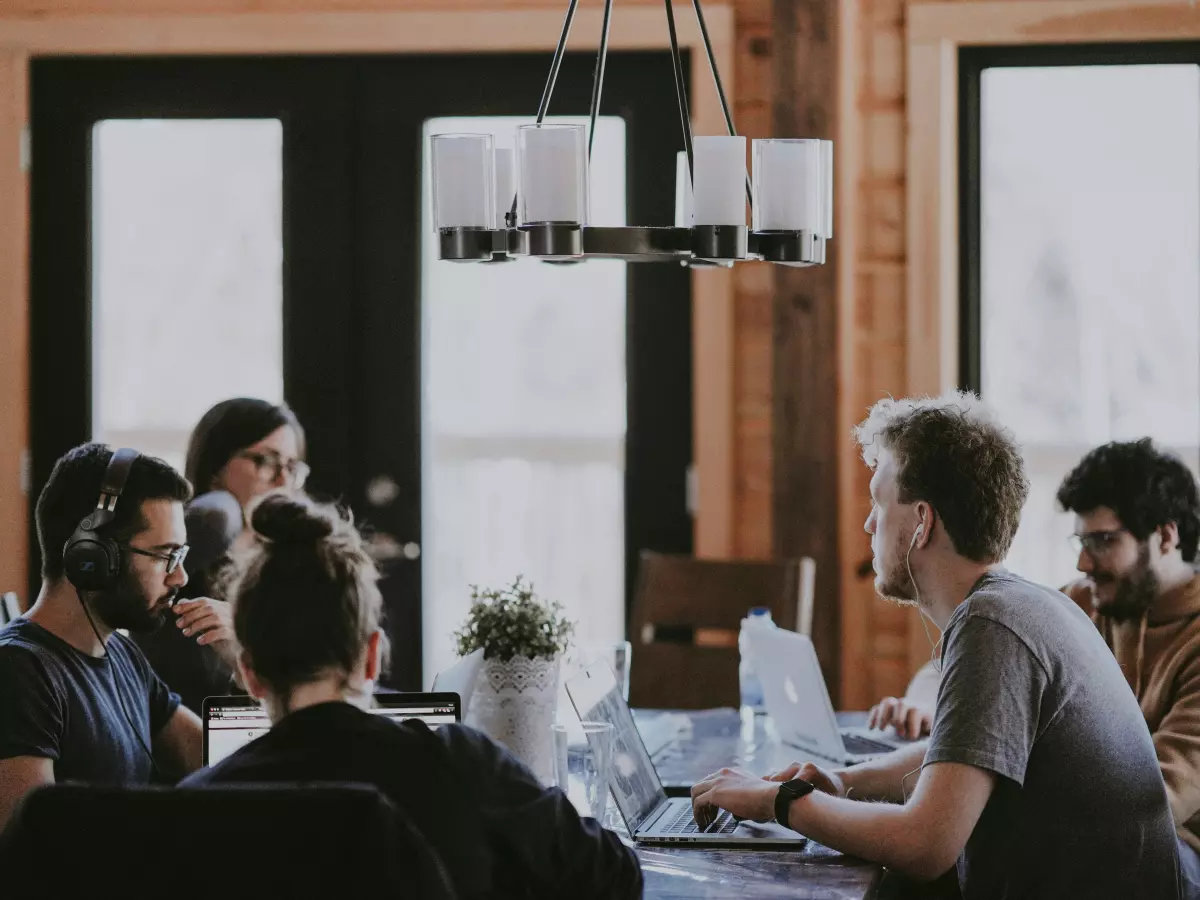 A group of young professionals collaborating around a large wooden table with laptops.