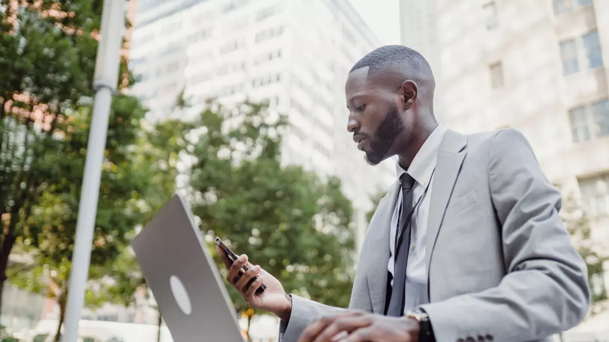 A man in a suit sitting on a bench in a city setting, using a laptop and a smartphone.