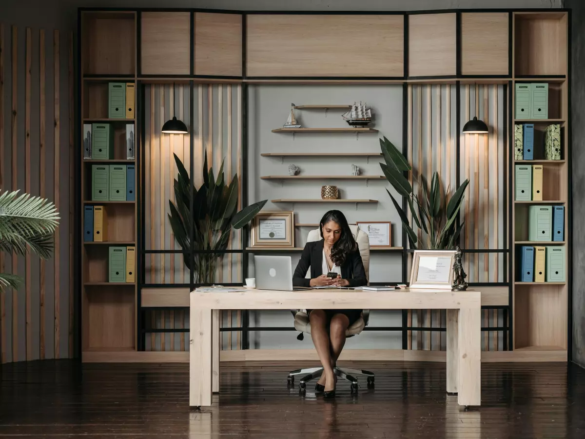 A woman in a suit sits at a large desk in a modern office with built-in bookshelves. The image emphasizes her central role and professional setting.