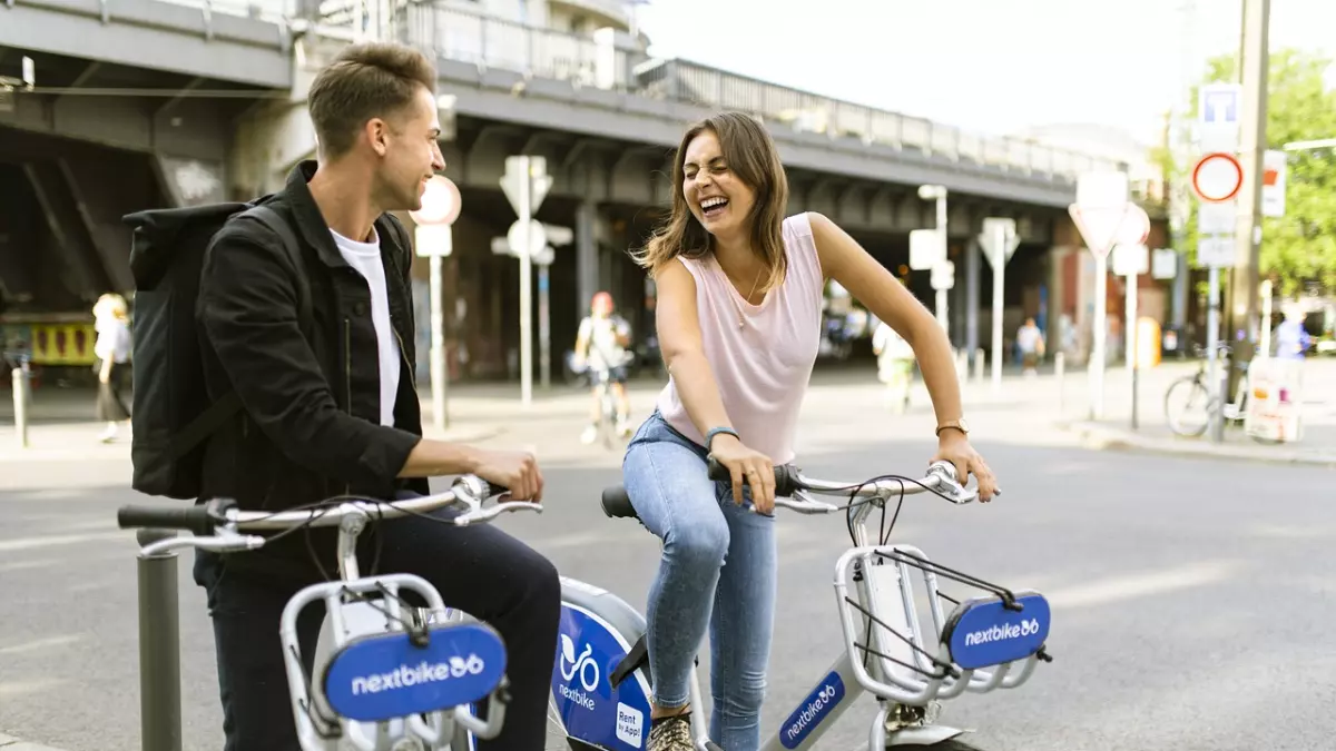 Two people on bikes are smiling and looking at each other, they are both holding smartphones.