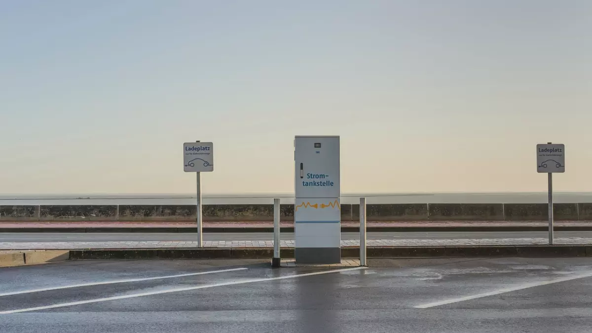 A charging station for electric vehicles is shown in the middle of a parking lot.  The station is a white box with a black sign that says "Strom-tankstelle." There are two signs on the left and right of the station that say "Ladeplatz."