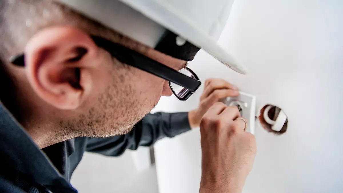 A man in a hard hat and glasses is working on an electrical outlet on a wall.