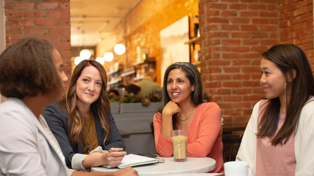 Four people, three women and one man, are sitting at a table in a cafe, talking and looking at each other. The cafe is decorated with exposed brick walls and warm lighting. The people appear to be casually dressed, and they have a relaxed and friendly atmosphere. The image is well-lit, and the composition is balanced.