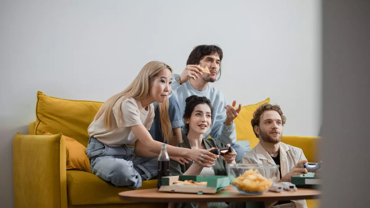 Four people playing a video game on a console, sitting on a yellow couch in a living room, surrounded by snacks and drinks.