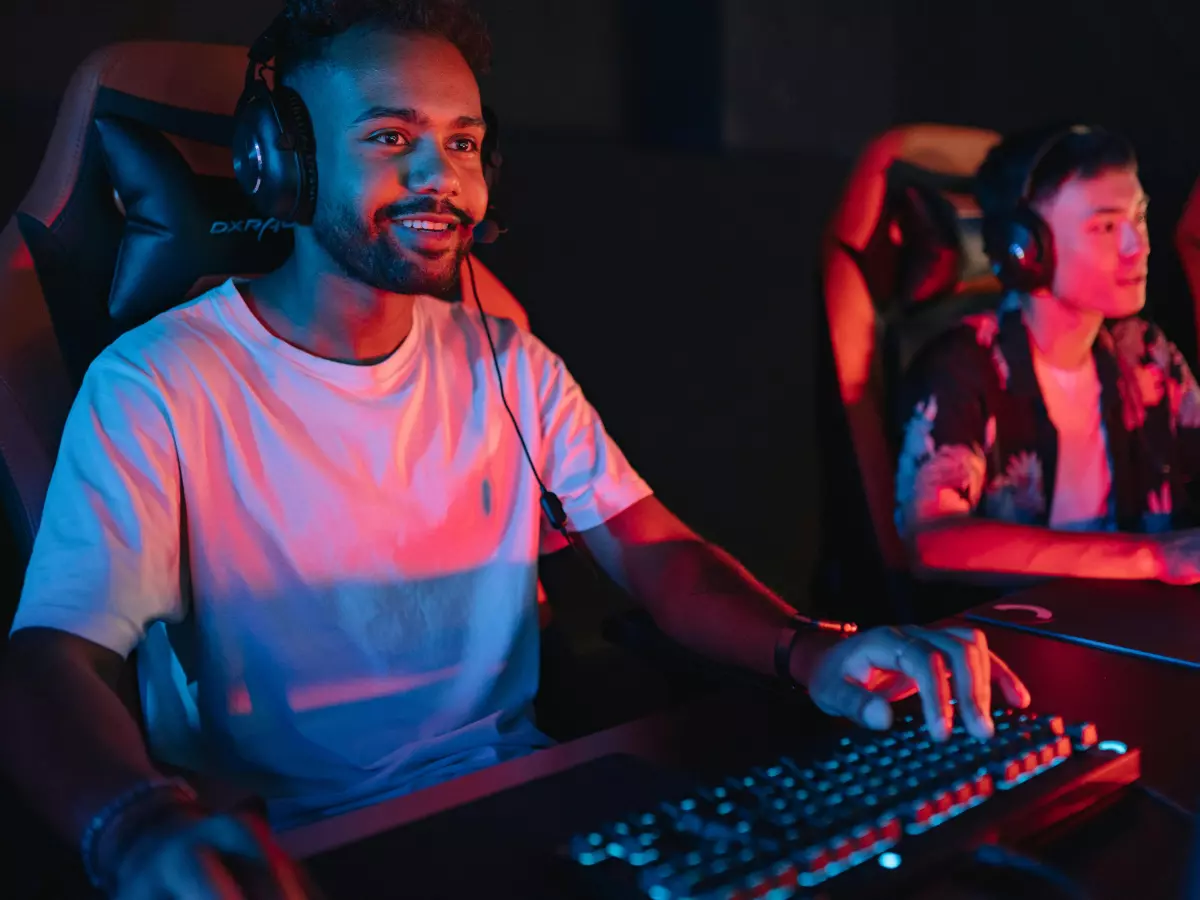 Two young adults wearing gaming headsets play video games in a dimly lit room.