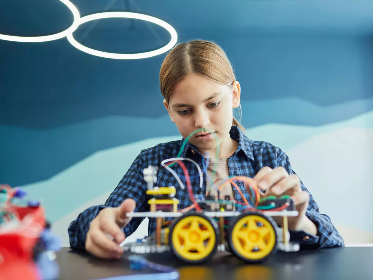 A young girl meticulously works on assembling a small robot, focusing intensely on the wiring.