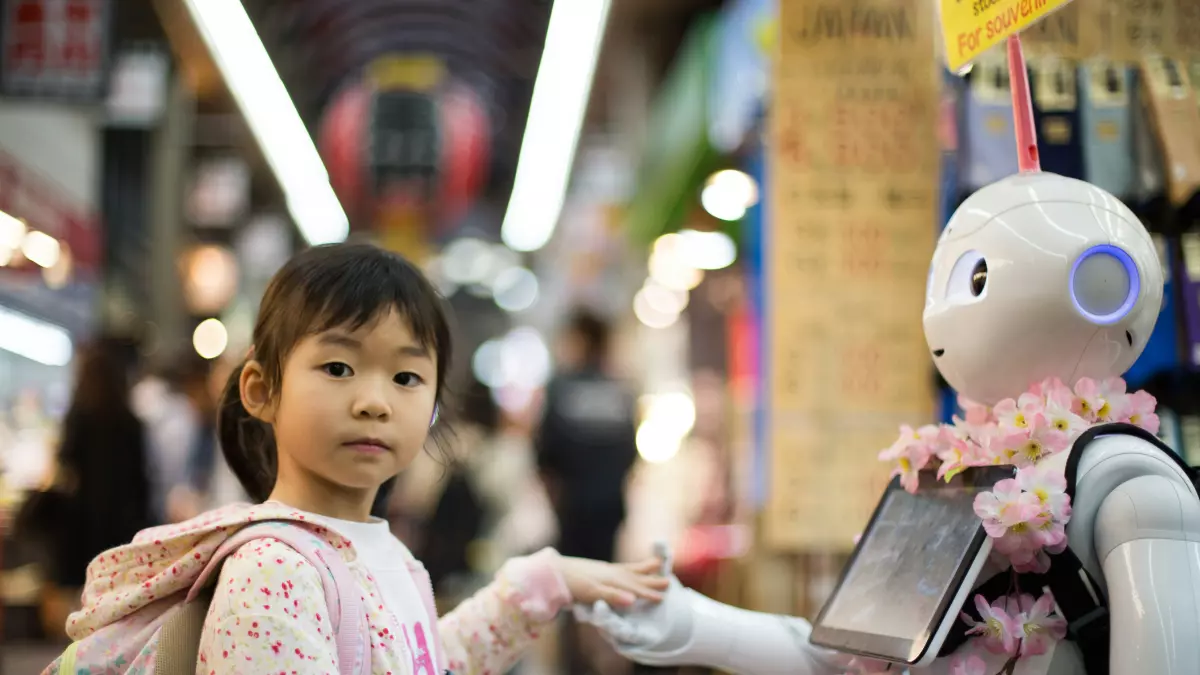 A young girl with a backpack stands facing a humanoid robot with a tablet. The robot is decorated with pink flowers and has a blue light around its eye.