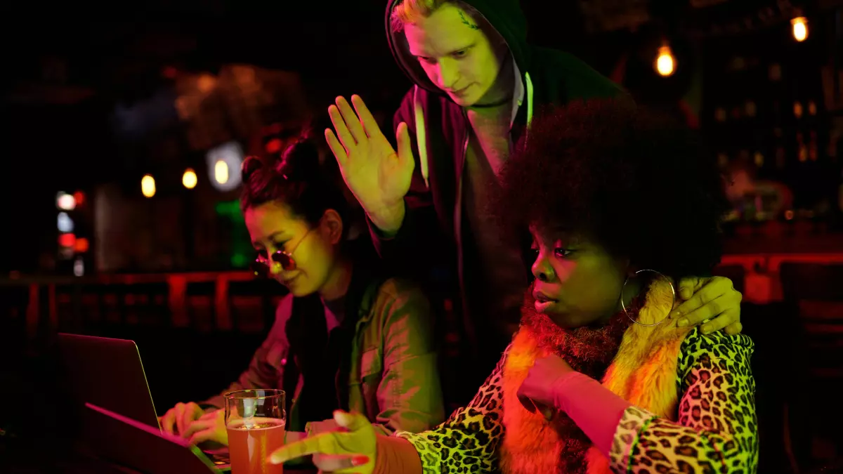 Three young women huddled around a laptop in a dimly lit bar setting. One woman gestures emphatically.