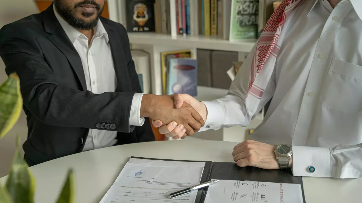 Two men in suits shaking hands at a table with documents and a pen on it.