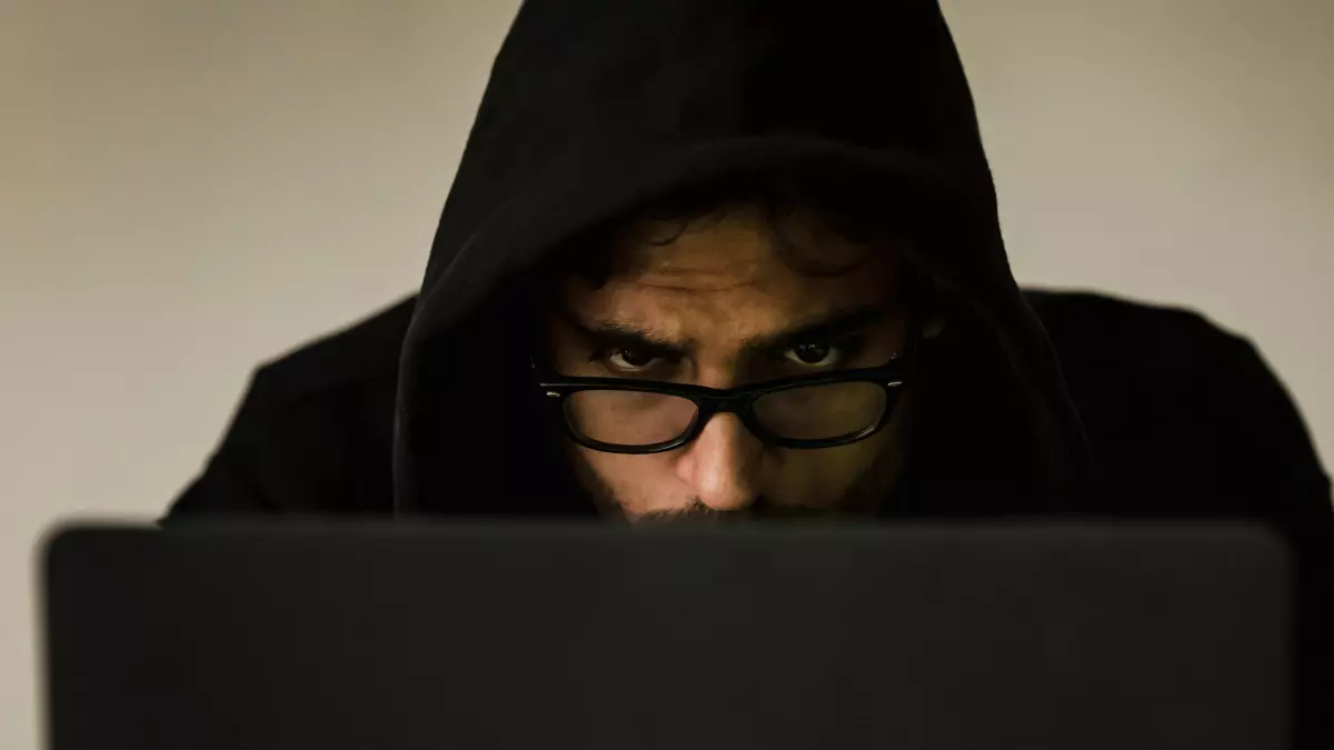 A close-up of a hooded figure peering over a laptop screen, with only their eyes visible. The image has a dark, mysterious atmosphere, suggesting the figure is involved in criminal activity.