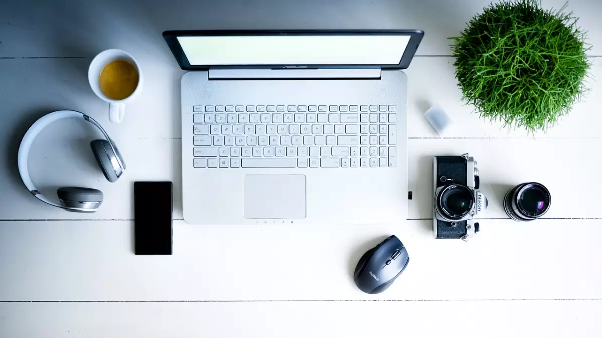 A white laptop, a smartphone, headphones, a camera, a mouse, a cup of coffee and a plant on a white wooden table.