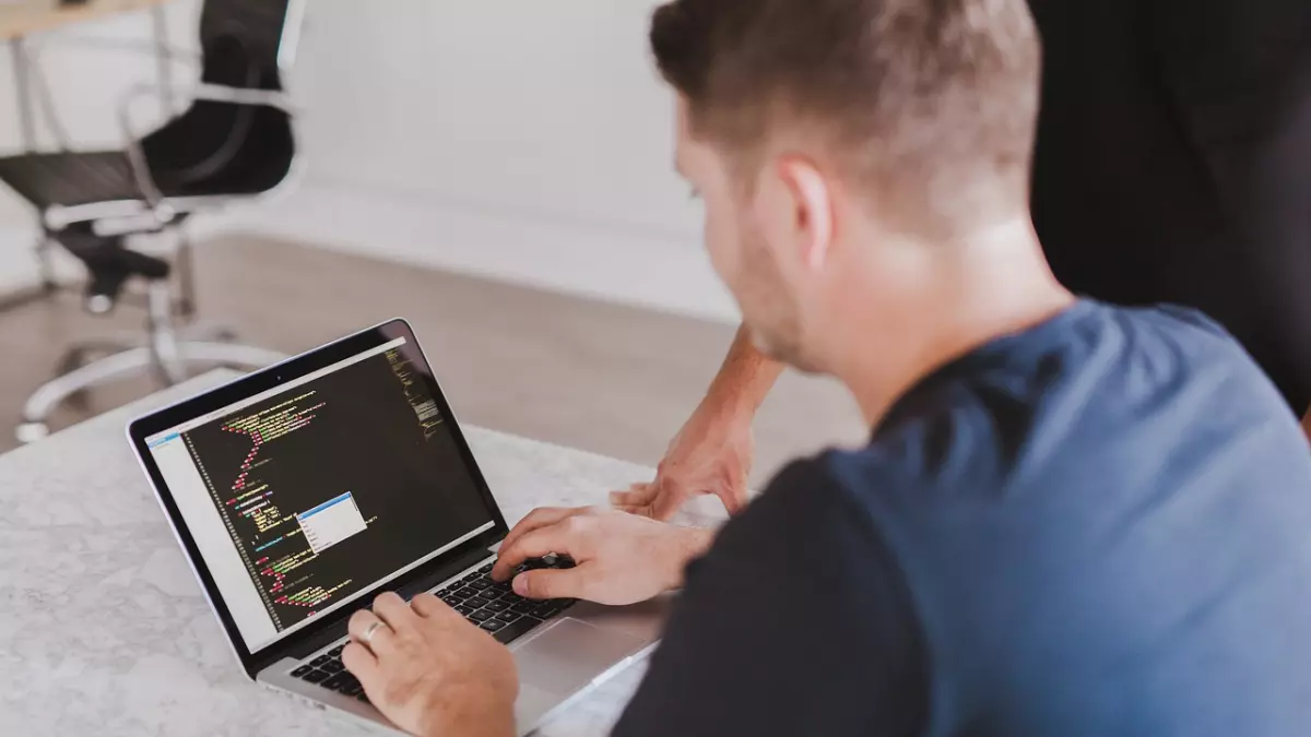 A person is sitting at a desk and typing on a laptop. The laptop screen shows code. The person is wearing a blue t-shirt and has their back to the camera.