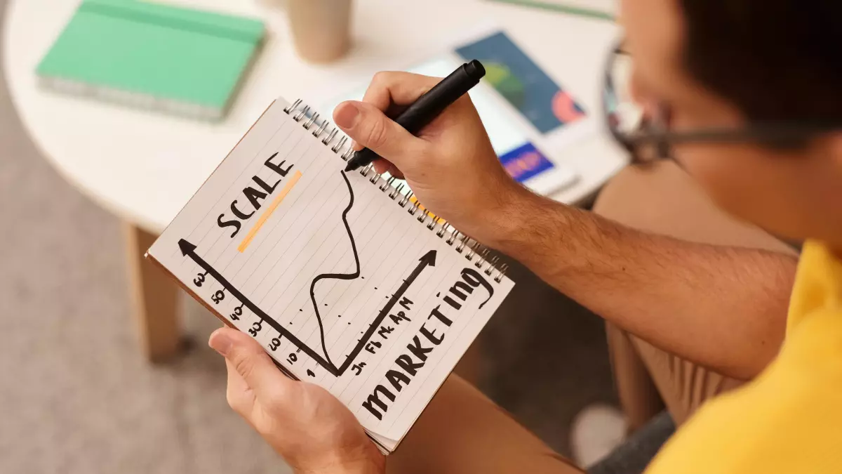 A person in a yellow shirt is writing "SCALE" on a notebook with a graph related to market growth, illustrating the importance of scaling in machine learning.