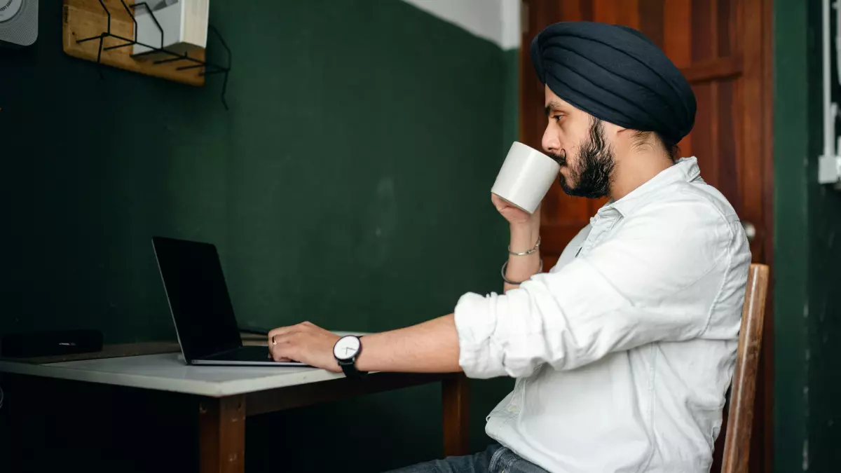 A man in a white shirt and blue turban is sitting at a desk, looking at a laptop. He is holding a white mug in his right hand. 