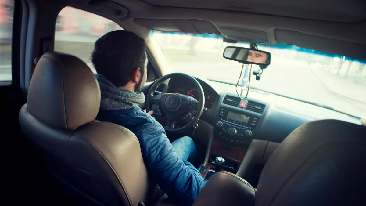 A man driving a car, seen from the rear passenger seat.