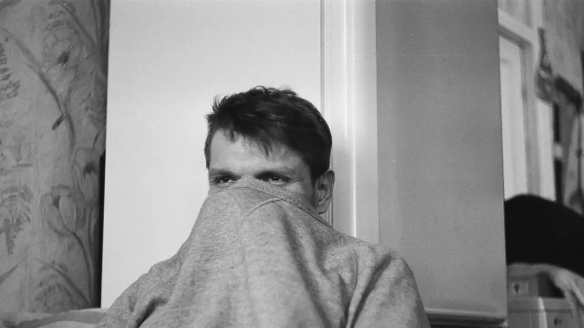 A black and white photo of a man hiding his face behind a blanket.