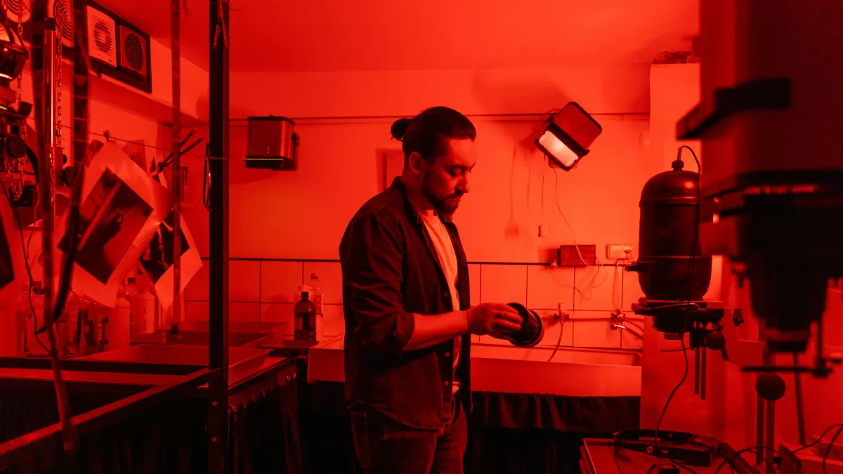 A man with a beard is standing in a room with red lighting, he is looking to the right and is wearing a black blazer and jeans. He is holding something in his right hand. There is a glass cabinet in the background, along with other objects.