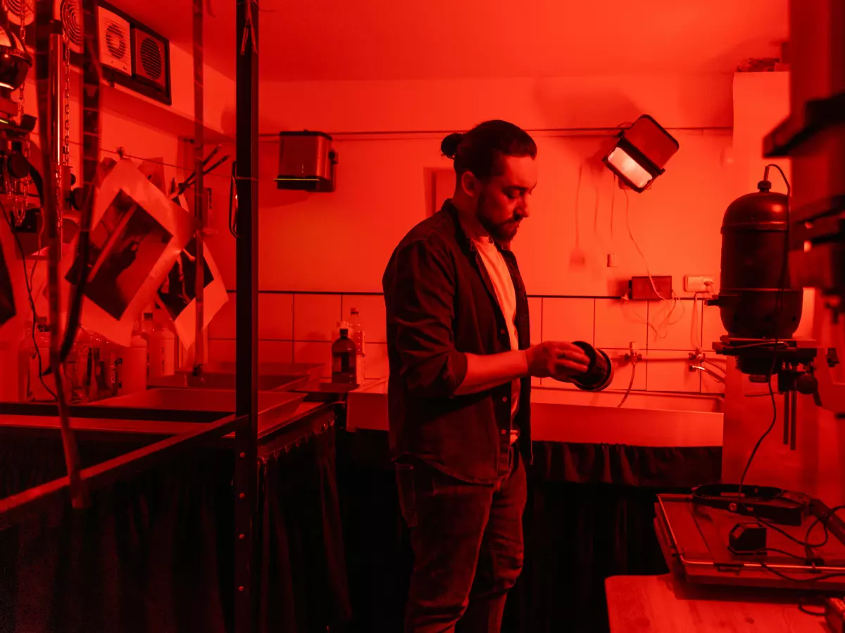 A man with a beard is standing in a room with red lighting, he is looking to the right and is wearing a black blazer and jeans. He is holding something in his right hand. There is a glass cabinet in the background, along with other objects.