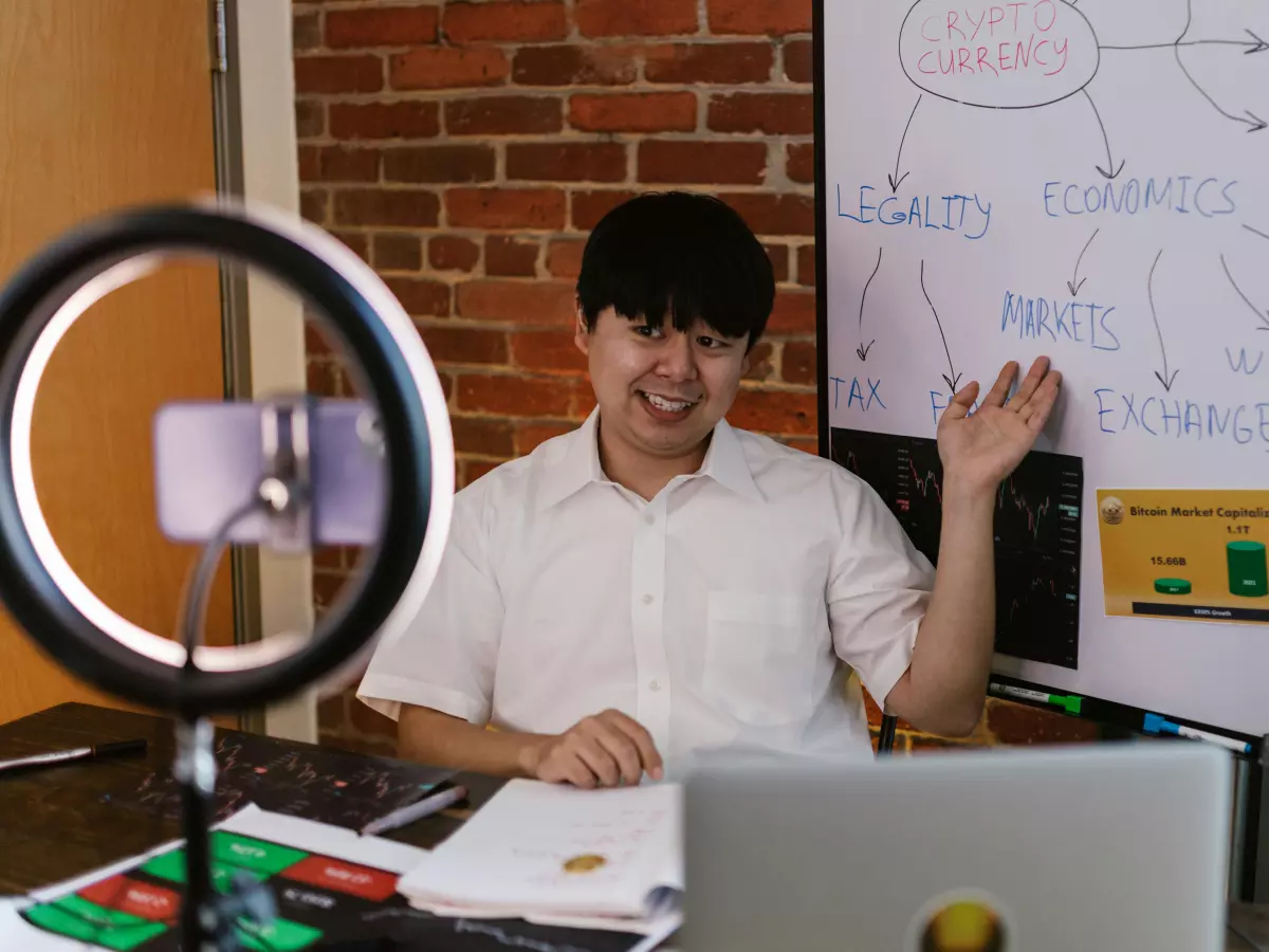 A man in a white shirt is sitting at a desk in front of a whiteboard with diagrams on it. He is pointing at the whiteboard with his right hand and smiling. There is a ring light and a microphone in front of him.