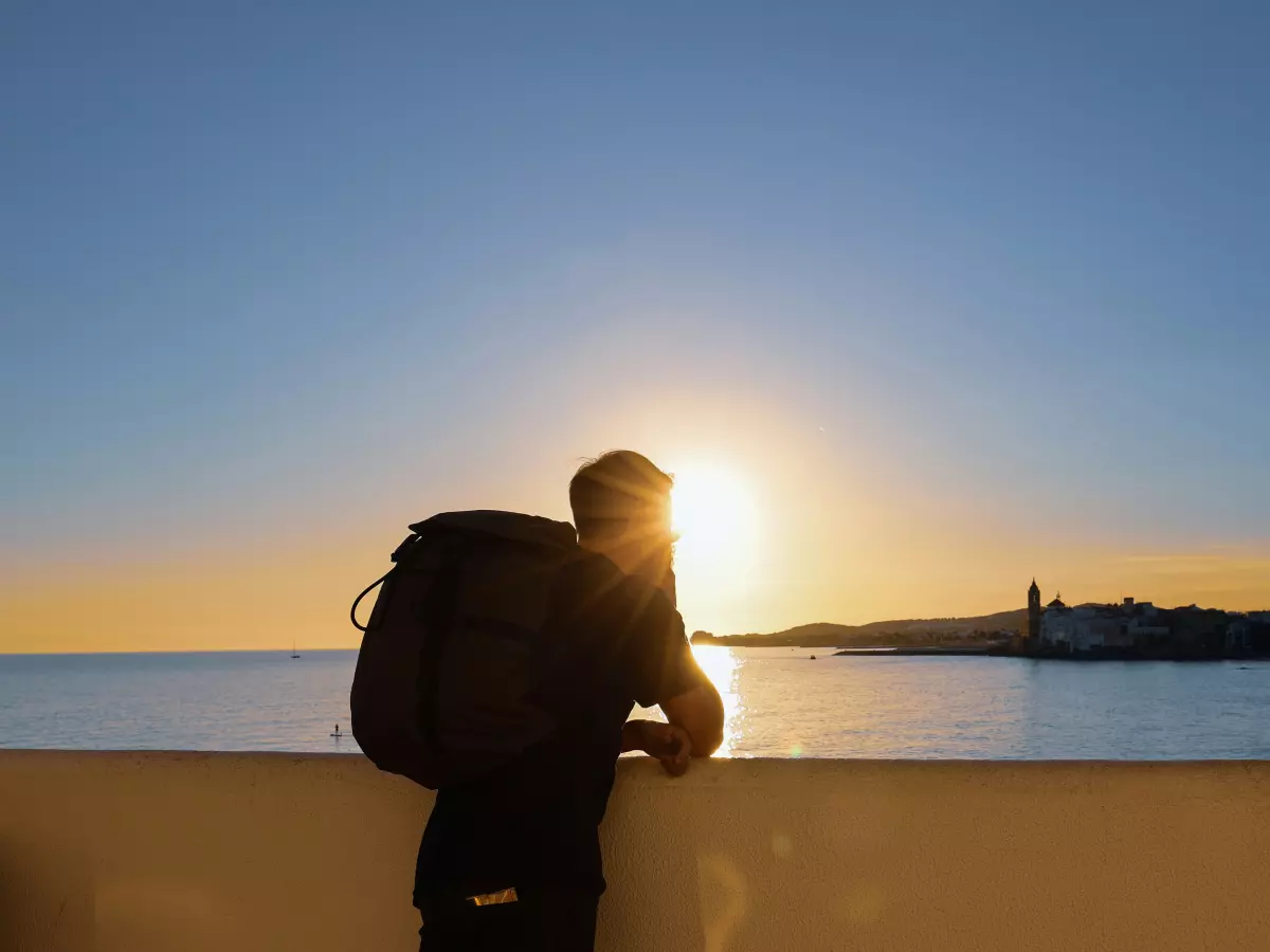 A person with a backpack standing on a wall overlooking the ocean with the sun setting in the background.