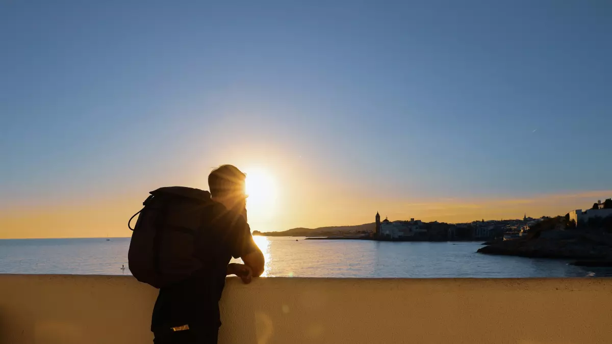 A person with a backpack standing on a wall overlooking the ocean with the sun setting in the background.