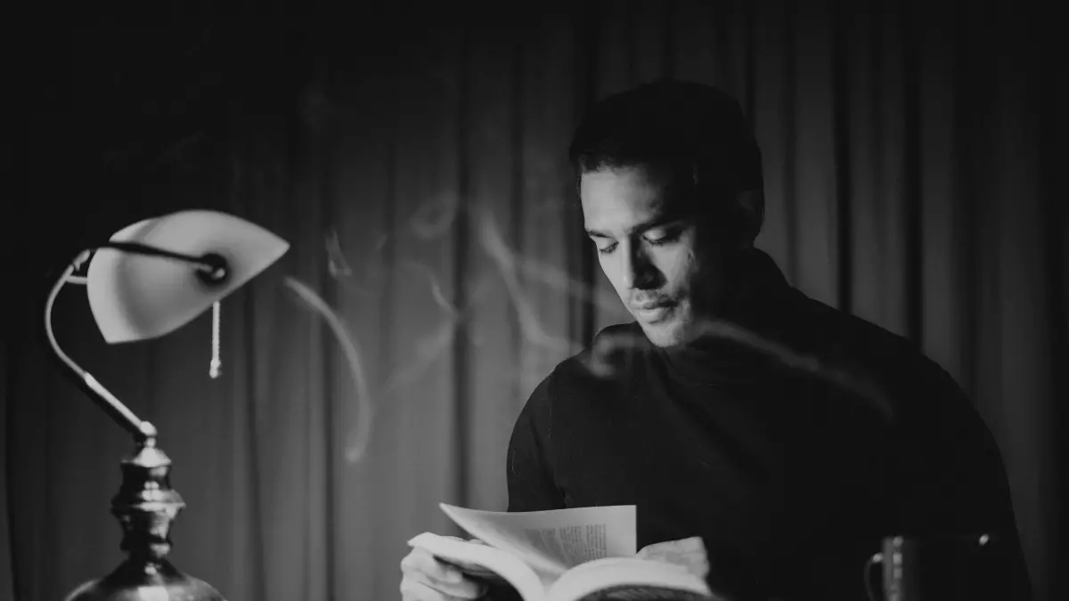 A man sitting at a desk in a dimly lit room, reading a book under a lamp. He is wearing a dark sweater and has a serious expression on his face.