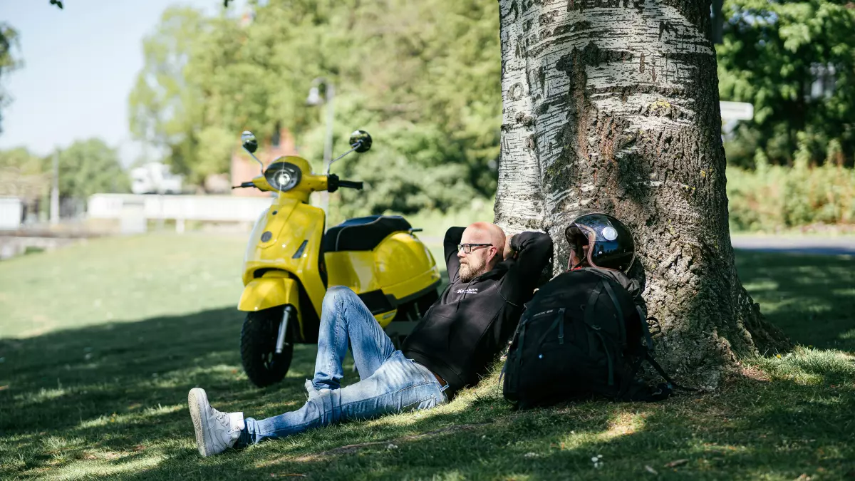 A man relaxing under a tree next to an electric scooter.