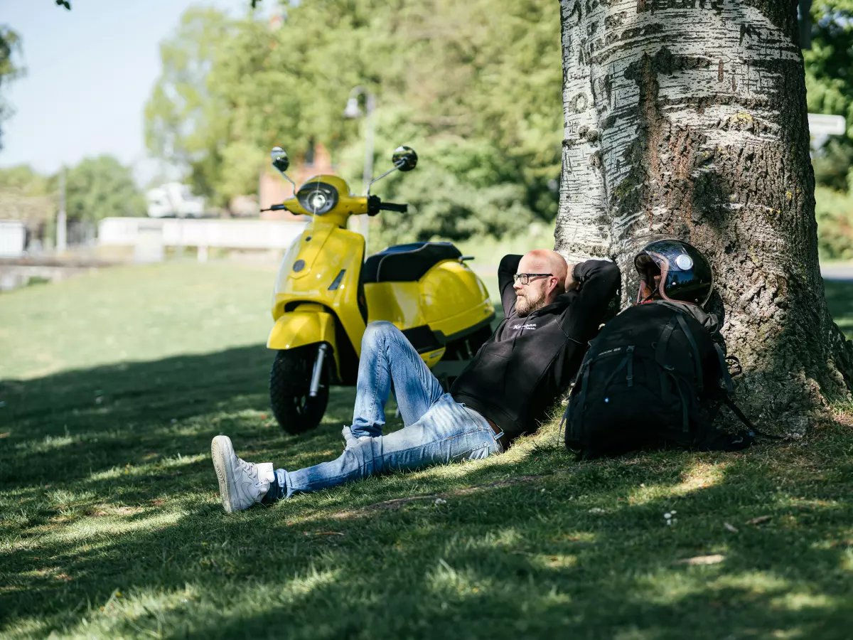 A man relaxing under a tree next to an electric scooter.