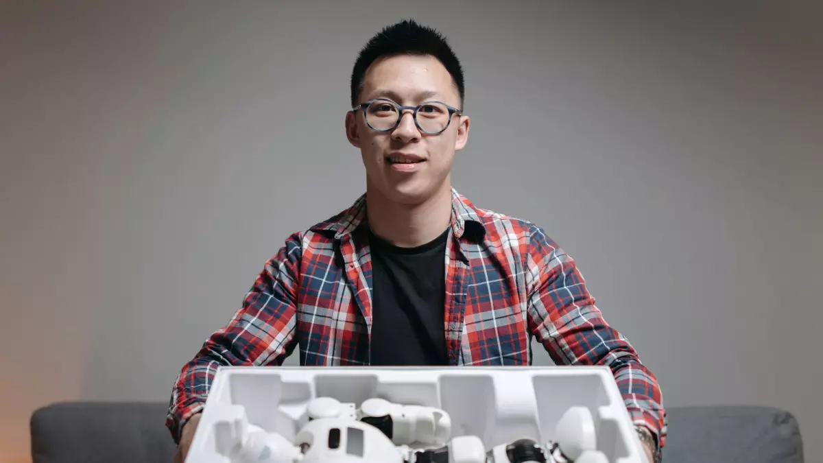 A man in a plaid shirt and glasses is unpacking a robot from its box, the robot is partially visible and the image is set in a living room.