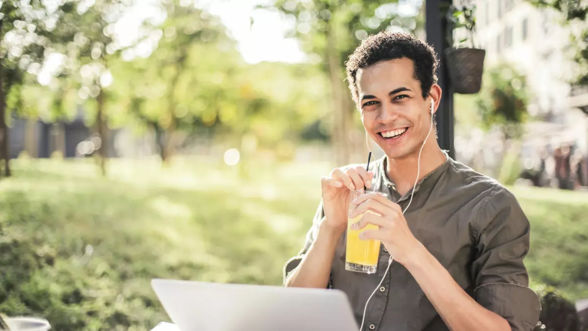 A young man sitting in a park, looking at the camera, drinking juice, with headphones on, while working on his laptop.