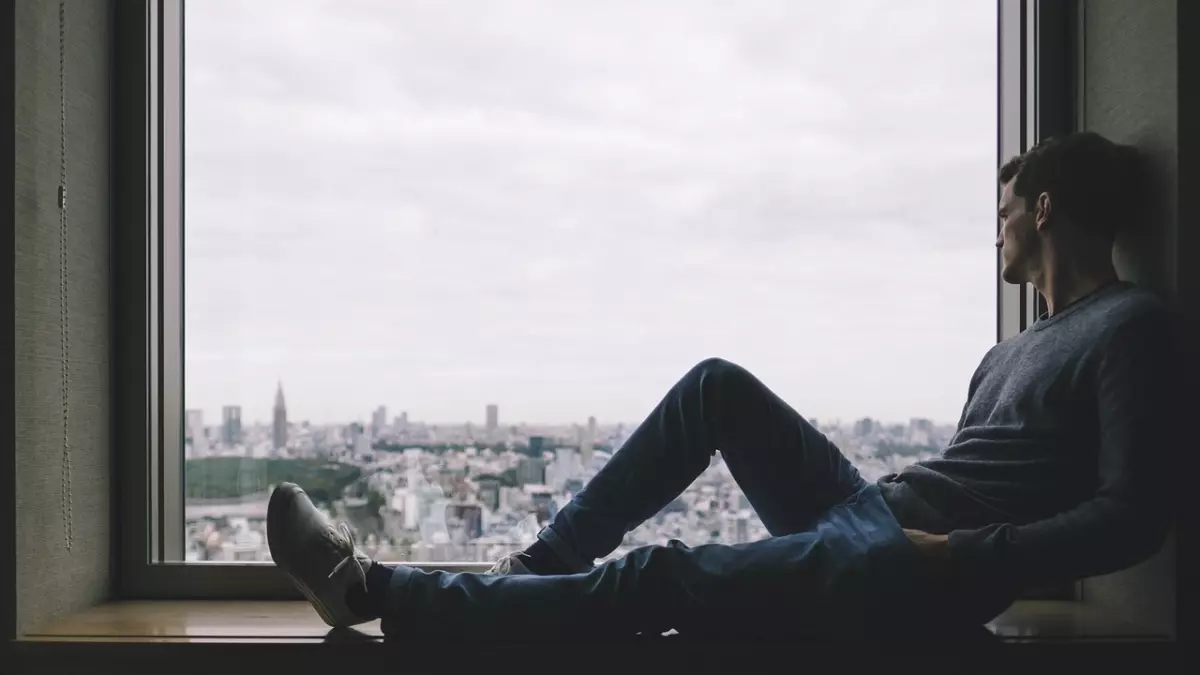 A man sits on a windowsill, looking out at a city skyline. He