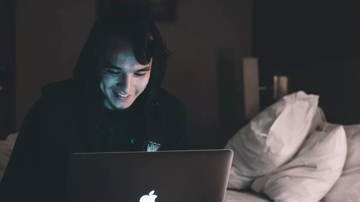 A young man is sitting in bed and looking at a laptop. He