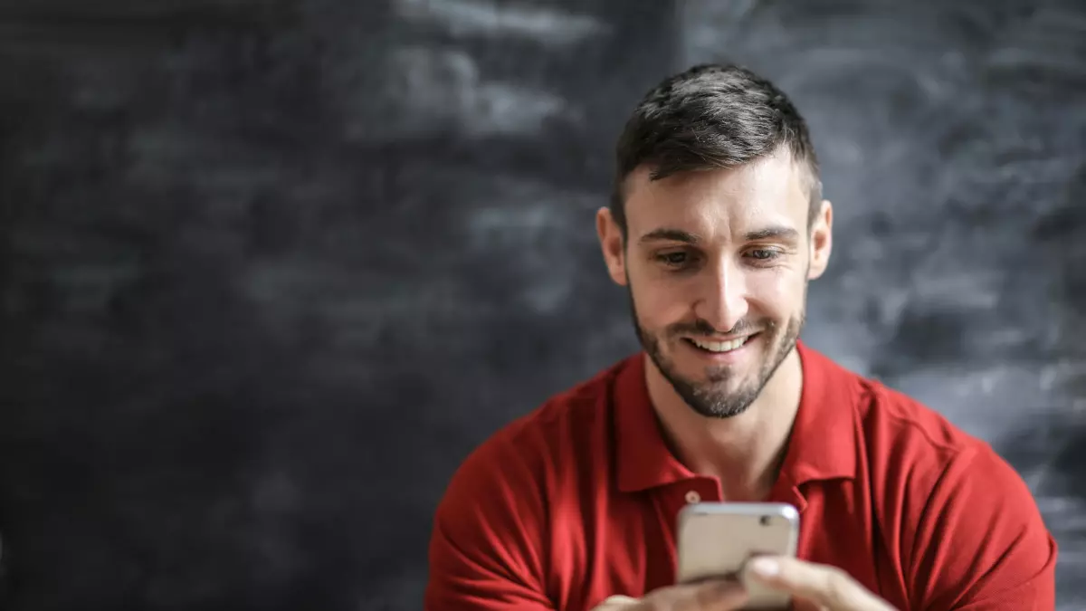 A young man sitting in a casual setting, smiling as he looks at his smartphone.