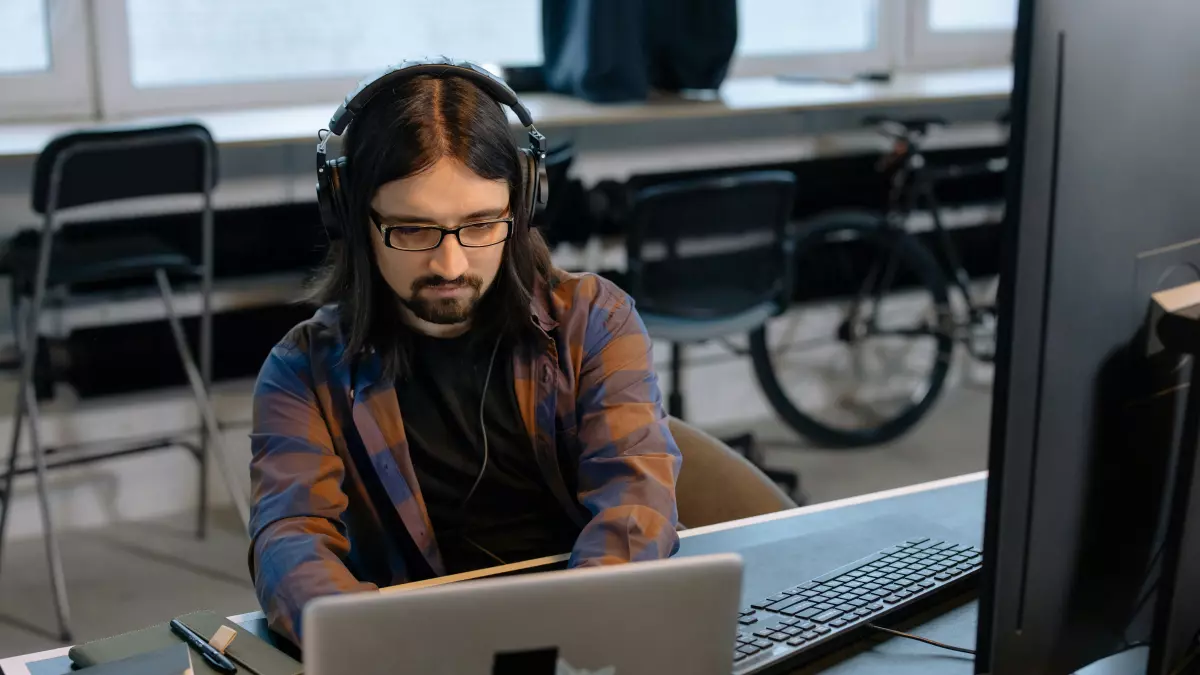 A man in a plaid shirt and headphones sits at a desk in front of two monitors, typing on a laptop. 