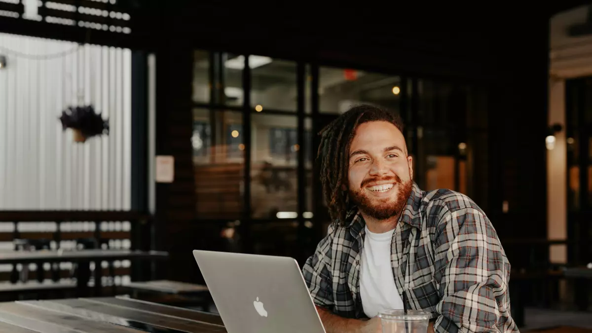 A man in a plaid shirt is sitting at a table with a laptop in front of him. He is smiling and looking to the side.