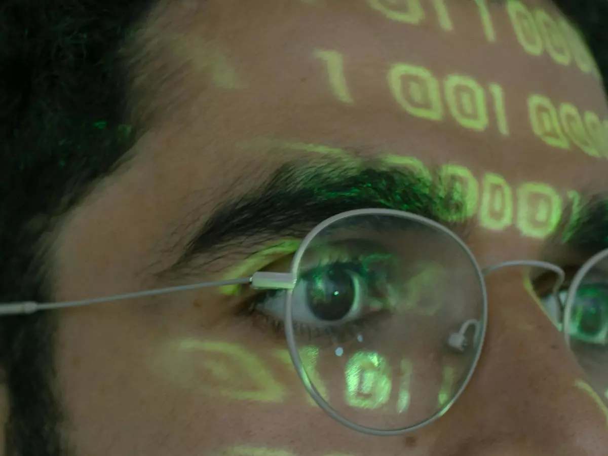 Close-up shot of a person's face with binary code projected onto their forehead and visible through their glasses. The image has a mysterious, tech-focused feel.