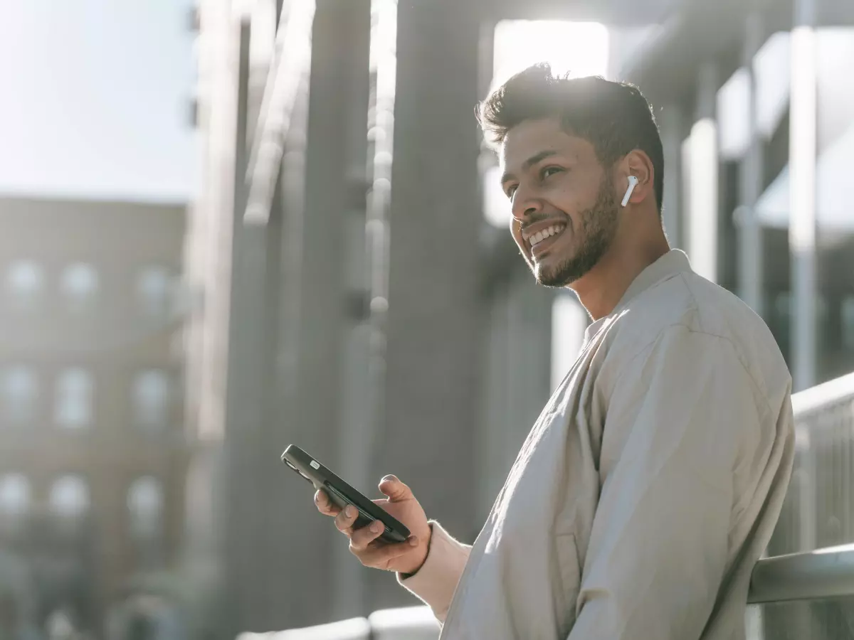 A young man with a beard is smiling and looking at his phone. He is wearing a light-colored shirt and standing outside with a city in the background.