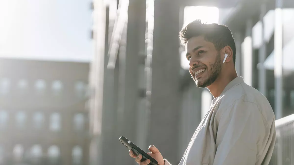 A young man with a beard is smiling and looking at his phone. He is wearing a light-colored shirt and standing outside with a city in the background.