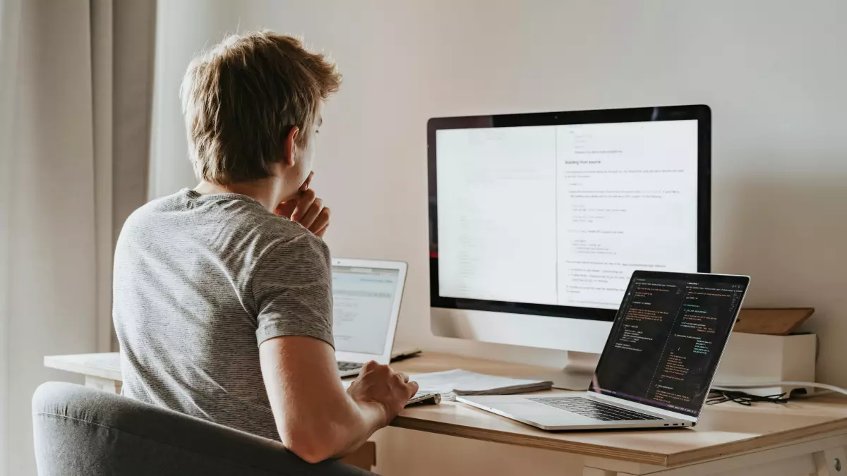 A young man sits at a desk facing two computer screens, one a large monitor displaying code. He is looking intently at the screen with his hand on his chin, appearing focused and contemplative. This image captures the essence of software development and the meticulousness required for effective code composition.