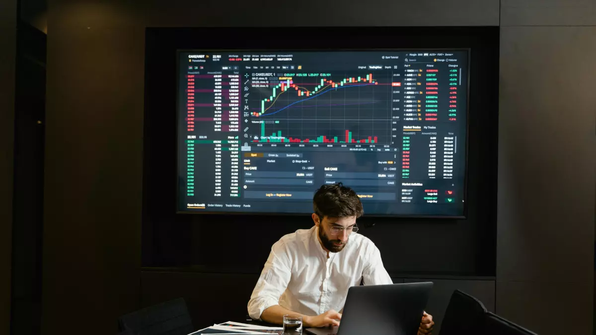 A man sitting at a desk, working on a laptop with a large screen displaying data charts behind him.