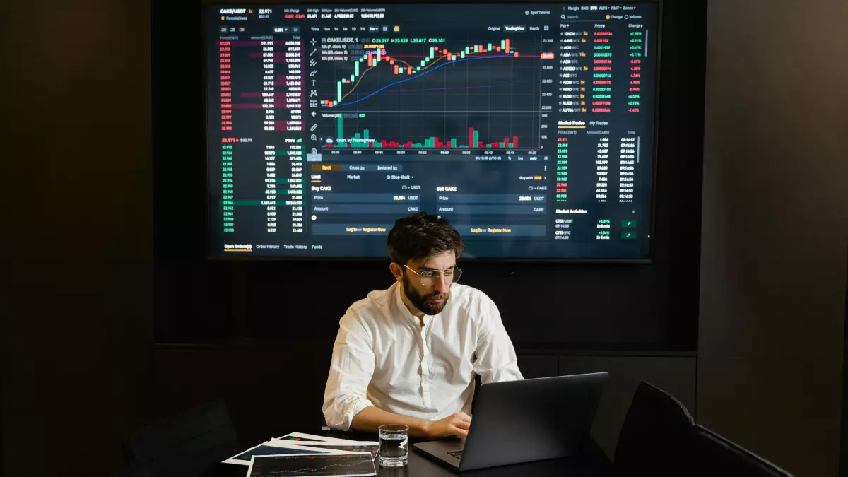 A man sits at a desk in front of a laptop, looking at a large screen with a data chart. He is wearing a white shirt and has a beard.