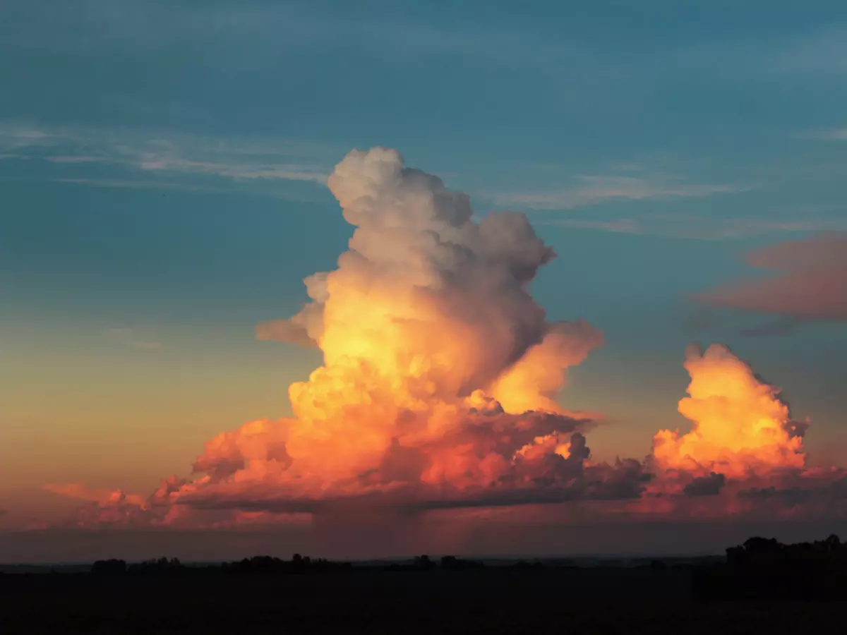 A dramatic sky with a large, puffy cumulus cloud illuminated by the setting sun, casting a warm orange glow. The cloud appears to be growing and expanding, with a smaller cloud to the right. The sky is a gradient of blue and orange, with a hint of pink.