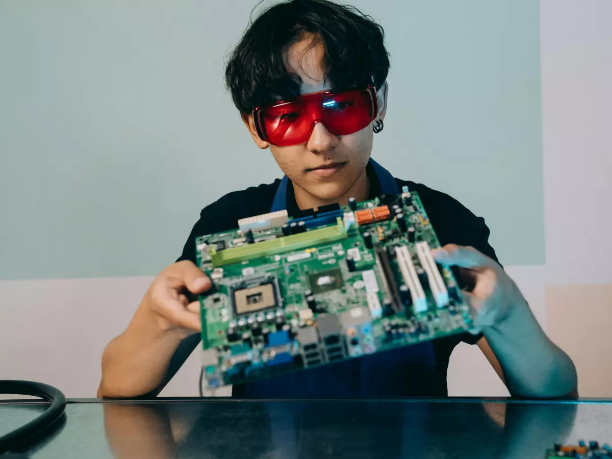 A person wearing red safety glasses holds a motherboard with a soldering iron on the table next to them.