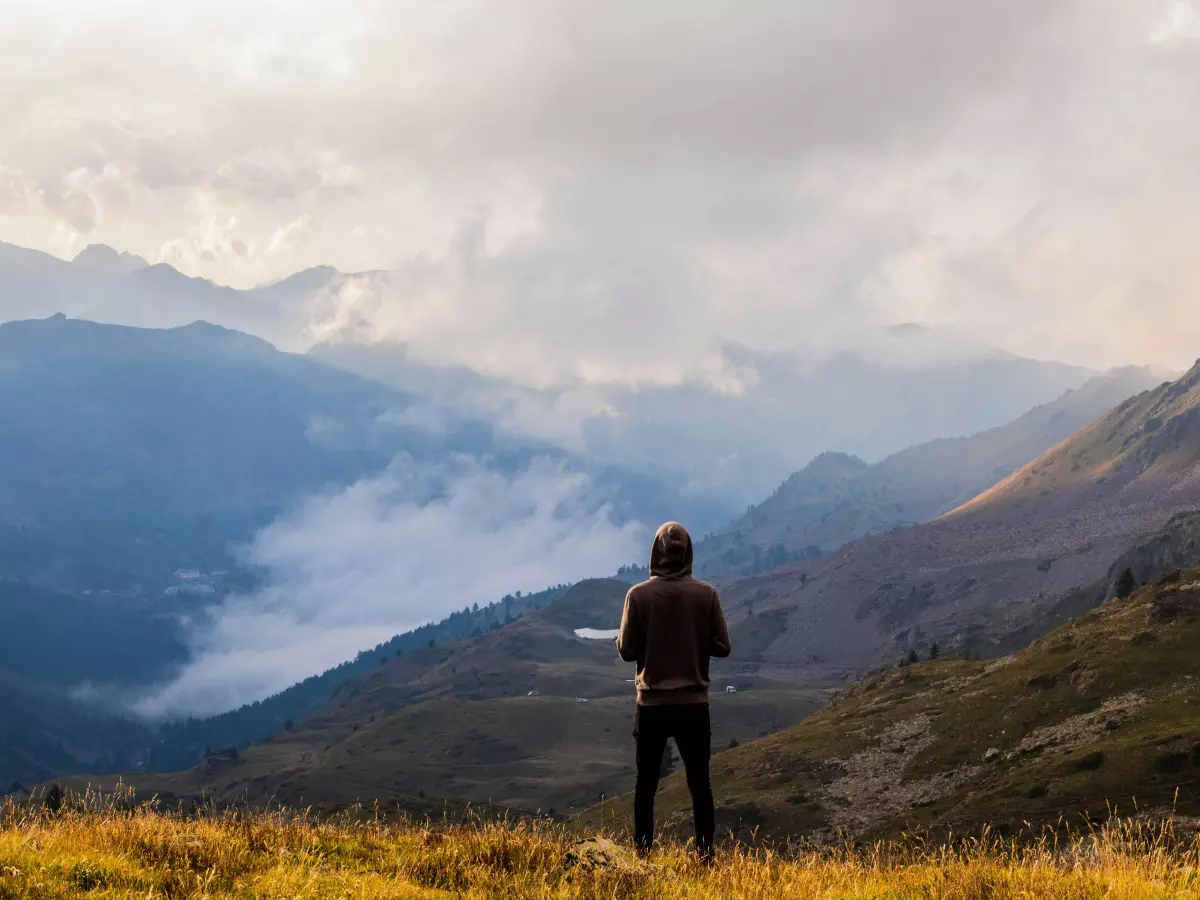 A man stands on a grassy hill, looking out at a vast, mountainous landscape. The mountains are covered in clouds, and the sky is blue.