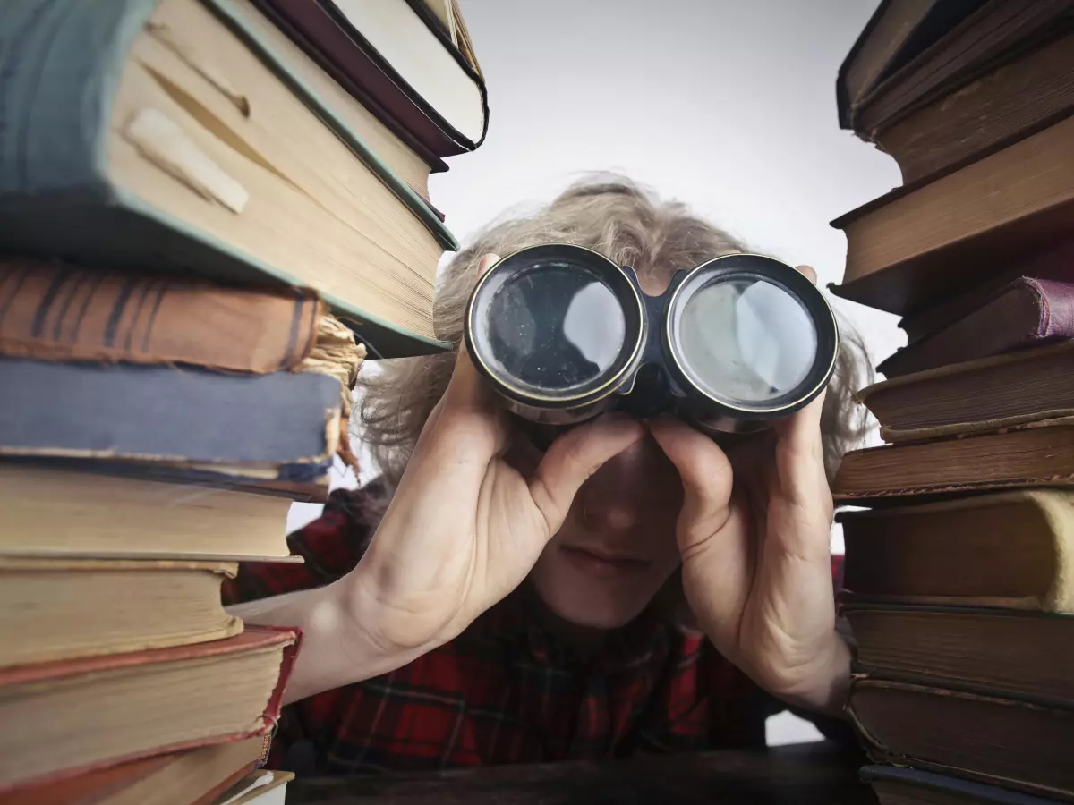 A person with blonde hair looks through binoculars, surrounded by a stack of books.